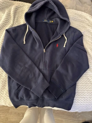 Marinblå hoodie från Polo Ralph Lauren - Snygg marinblå hoodie från Polo Ralph Lauren med dragkedja, vit dragsko i huvan och den klassiska röda loggan broderad på bröstet.
