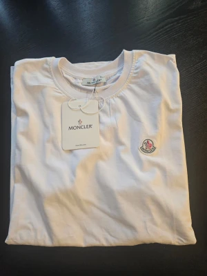 Vit Moncler t-shirt med logga - Snygg vit t-shirt från Moncler med klassisk rund halsringning och broderad logotyp på bröstet. Tillverkad i mjuk bomull för en skön känsla. Perfekt för dig som gillar stilrena och exklusiva plagg. Strl. XXL helt ny med prislappar kvar 