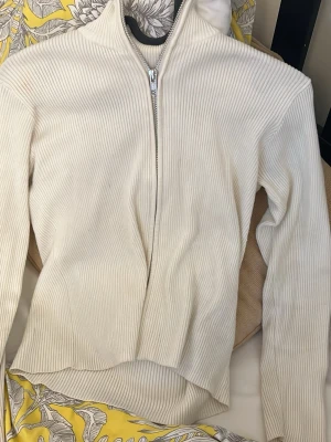 Vit ribbad ziptröja med hög krage - Säljer en stilren vit ribbad ziptröja med hög krage. Tröjan har långa ärmar och dragkedja framtill. Materialet är stretchigt och följsamt, perfekt för lager på lager. Enkel och clean design som passar till det mesta.