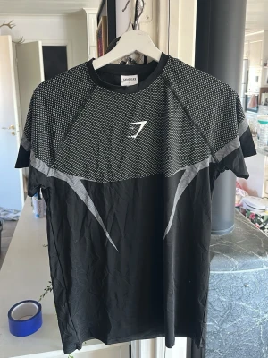 Svart träningströja från Gymshark - Snygg svart träningströja från Gymshark med grått mönster över axlar och bröst samt logga framtill. Tillverkad i ett lätt och stretchigt material som andas, perfekt för gymmet eller löpning. Kortärmad och med modern passform. OBS rep, passar S som muselfit och passar även M fast lite tajtare 