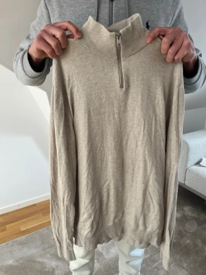 Beige halvzip-tröja från Jack & Jones - Snygg beige tröja från Jack & Jones med halv dragkedja och hög krage. Tillverkad i mjukt material och har ribbade muddar vid ärmslut och nederkant. Perfekt för en clean och stilren look.