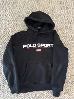 Svart hoodie Polo Sport Ralph Lauren - Svart hoodie från Polo Sport Ralph Lauren med stor vit logga och röd text på bröstet samt en liten amerikansk flagga. Klassisk känguruficka framtill och justerbar huva med snören. Tillverkad i mjukt bomullsmaterial för skön känsla. Mycket bra skick, använt ca 4-5 gånger, säljer då den inte kommer till användning 