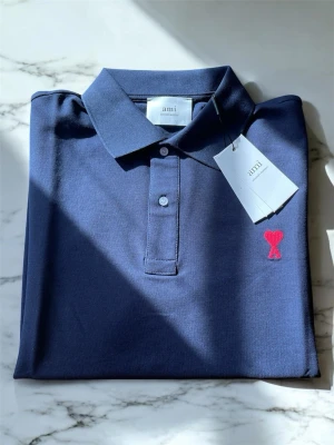 Mörkblå pikétröja från Ami - Snygg mörkblå pikétröja från Ami med klassisk krage och knappar framtill. Tröjan har korta ärmar och ett rött hjärtemblem broderat på bröstet. Tillverkad i mjuk bomull som känns skön mot huden. Perfekt för en clean och stilren look.