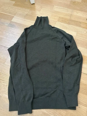 Mörkgrön polotröja stickad stl M - Mörkgrön stickad polotröja med lång ärm och ribbade muddar vid ärmslut och nederkant. Tröjan har en hög polokrage och är tillverkad i ett mjukt, tunt material som passar perfekt för lager på lager. Enkel och stilren design utan synliga loggor.