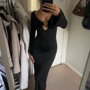 Svart långklänning med guldig detalj - Supersnygg svart långklänning med djup v-ringning och lång ärm. Klänningen har en unik cut-out framtill med en guldig blomdetalj. . Materialet är stretchigt och sitter tight, med lätt utsvängda ärmslut. Aldrig använd! 