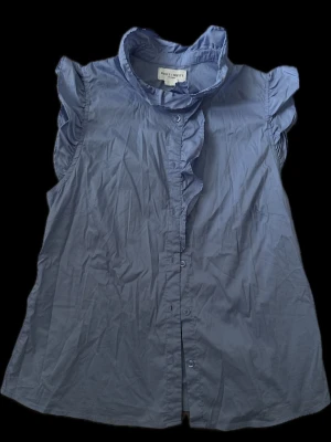  ärmlös blus med volang - Snygg blå blus från Holly & Whyte med volangdetalj längs knäppningen och liten ståkrage. Blusen är ärmlös med lätt puffade axlar och har en rak passform Inga defekter.