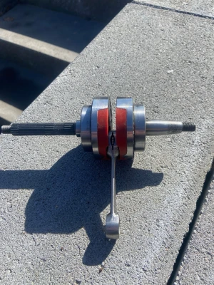 Röd och silver vevaxel till moped - Säljer en vevaxel i metall med röda detaljer, perfekt för dig som bygger eller lagar moped. Vevaxeln har två axlar med gängor och en kopplingsarm. Passar till flera olika mopeder och är tillverkad i slitstarkt material.