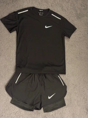 Nike miller - Säljer ett svart träningsset från Nike med t-shirt och shorts. Båda plaggen har vita Nike-loggor och reflekterande detaljer. Materialet är lätt och andas, perfekt för löpning eller gym. T-shirten har rund hals och shortsen har elastisk midja.