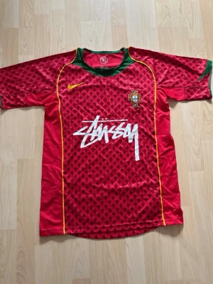 Röd Stussy x Nike Portugal T-shirt - Unik röd t-shirt med Portugal-emblem, Stussy-logga i vitt och Nike swoosh i gult. T-shirten har gröna detaljer vid kragen och ett mönster över hela framsidan. Perfekt för dig som gillar streetwear och fotbollsinspirerad stil.