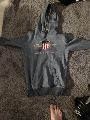 Grå hoodie från GANT med tryck - Snygg grå hoodie från GANT med stort tryck och logga på bröstet samt texten 'American Sportswear'. Klassisk känguruficka framtill och justerbar huva med snören. Perfekt för en avslappnad och sportig stil.