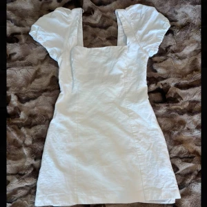 Vit kortklänning med puffärmar och snörning - Superfin vit kortklänning med fyrkantig ringning, puffiga kortärmar och snörning i ryggen. Klänningen har dragkedja i sidan och är gjord i ett lätt material, perfekt för sommaren! På första bilden kan man se att dragkedjan inte går att dra upp hela vägen på grund av lösa stygn ( 3:e bilden) annars i bra skick!🥰