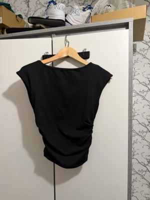 Svart ärmlös topp med drapering - Svart topp med draperad effekt på sidan och breda axlar. Modellen är ärmlös och har en enkel, stilren design som passar till många olika outfits. Materialet känns mjukt och stretchigt, perfekt för att skapa en snygg siluett.