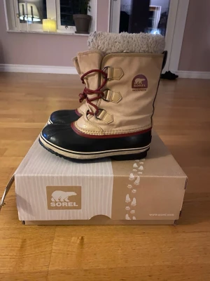 Beige vinterstövlar från Sorel - Helt oanvända beige vinterstövlar från Sorel med svart gummisula, röda snören och varm, fluffig insida. Vattentäta och perfekta för kalla dagar. Klassisk design med kontrastfärgade detaljer och robust yttersula för bra grepp.
