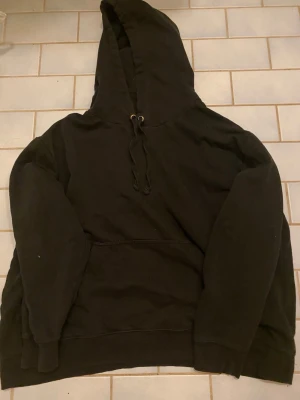 Svart hoodie med huva och magficka - En klassisk svart hoodie med stor huva och dragsnören. Hoodien har en rymlig magficka framtill och ribbade muddar vid ärmslut och nederkant. Perfekt för en avslappnad och street-inspirerad stil. Materialet känns mjukt och bekvämt.