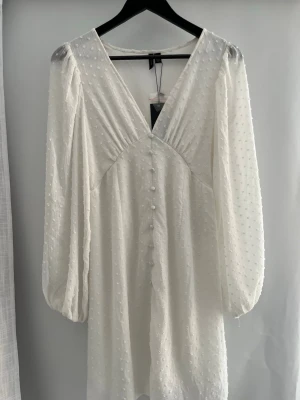Vit prickig klänning med knappar - Säljer en vit klänning med diskreta prickar i tyget, v-ringning och klädda knappar framtill och ett resårband baktill i midjan. Klänningen har långa, puffiga ärmar och är gjord i ett lätt, skönt material. Den är ny med prislapp kvar. Perfekt som student eller sommarklänning💕