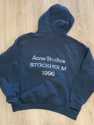 Mörkblå hoodie Acne Studios - Mörkblå hoodie från Acne Studios med stor huva och tryck på ryggen där det står 'Acne Studios STOCKHOLM 1996'. Hoodien har ribbade muddar vid ärmslut och nederkant. Tillverkad i mjukt bomullsmaterial för en avslappnad och skön look.