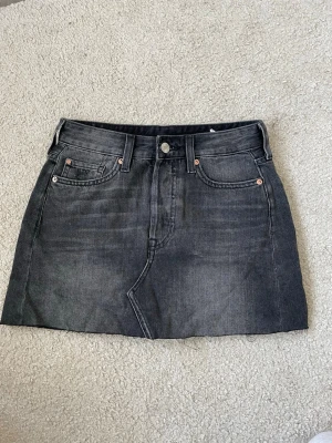 Svart jeanskjol med fem fickor - Snygg svart jeanskjol i kort modell med klassisk femficksdesign och knappgylf. Kjolen har en lätt tvättad look och raka linjer, perfekt till vardags eller när du vill ha en chill outfit.