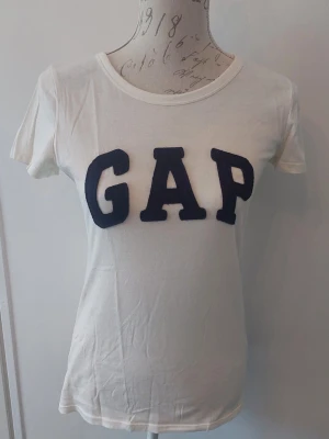 Vit GAP t-shirt med tryck - Vit t-shirt från GAP med stora mörkblå bokstäver på bröstet. Klassisk rund halsringning och korta ärmar. Tillverkad i mjuk bomull för en skön känsla och enkel stil.