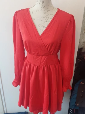 Röd omlottklänning med v-ringning - Snygg röd kortklänning med omlott och v-ringad hals. Klänningen har långa ärmar med smock vid ärmslut och markerad midja som ger en fin siluett. 