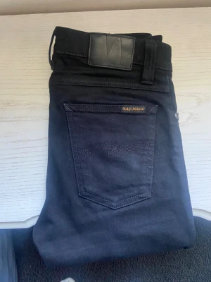 Svarta jeans från Nudie Jeans - Snygga svarta jeans från Nudie Jeans med klassisk femficksmodell och diskret logga på bakfickan. Jeansen har raka ben och är tillverkade i mjuk denim med en stilren look. Perfekta för dig som gillar minimalistisk och tidlös stil. 