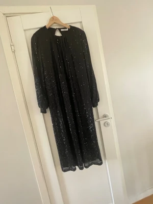 Svart glittrig långklänning med öppen rygg - Säljer en svart långklänning med glittriga detaljer och långärmad design. Klänningen har en snygg öppen rygg och ett lätt plisserat tyg som ger ett elegant fall. Perfekt för dig som vill sticka ut på festen!