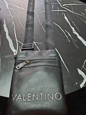 Svart axelväska från Valentino - Snygg svart axelväska från Valentino i slätt konstläder. Väskan har justerbar axelrem, två dragkedjefack och tydlig logga framtill. Perfekt för att bära det viktigaste och ger en clean look till din outfit.