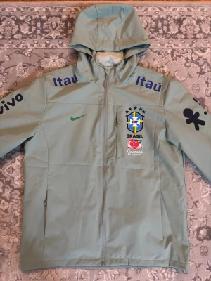 Nike Brasil vindjacka med huva - Ljusgrön vindjacka från Nike med huva och dragkedja. Jackan har Brasilien CBF-logga, sponsortryck och Nike-logga på bröstet. Flera tryck i blått och svart på ärmar och axlar. Perfekt för dig som gillar fotboll och sportig stil. PASSAR ÄVEN L