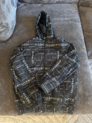 Svart vindjacka från Givenchy - Svart vindjacka med heltryckt Givenchy-logga i vitt över hela jackan. Jackan har huva, dragkedja framtill och elastiska muddar vid ärmslut och nederkant. Perfekt för dig som vill sticka ut med en streetstyle-look.