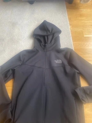  The North Face Hoodie  - Snygg svart hoodie från The North Face med dragkedja hela vägen och huva. Tillverkad i ett mjukt och stretchigt material, perfekt för chill eller träning. Logga på bröstet och ryggen. Klassisk och stilren design.