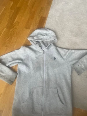 Grå  Polo Ralph Lauren zip hoodie - Snygg gråmelerad hoodie med dragkedja från Polo Ralph Lauren. Klassisk huva, två stora fickor fram och ikonisk mörkblå polospelare broderad på bröstet. Perfekt för en chill och stilren look. Mjuk bomullsblandning som känns skön mot huden.