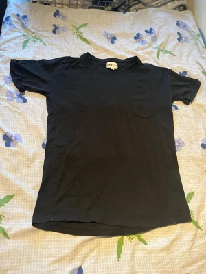 Svart basic t-shirt från AMI - En enkel svart t-shirt från AMI med rund hals och korta ärmar. Tillverkad i mjuk bomull och har en liten bröstficka på vänster sida. Perfekt till jeans eller shorts för en clean och avslappnad stil.