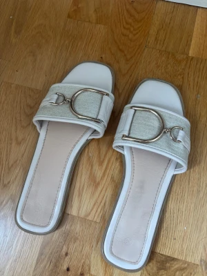 Vita sandaler med gulddetalj - Stilrena vita sandaler med fyrkantig tå och bred rem i textil och skinnimitation. Dekorerade med en stor guldfärgad metallspänne framtill. Snygg och modern design som passar till många outfits.