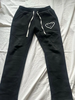 Carsicko joggers  - Säljer ett par svarta mjukisbyxor med vit dragsko i midjan och en broderad hjärtdetalj med smiley på ena benet. Byxorna har raka ben och två sidofickor. Perfekta för chill dagar och riktigt sköna att ha på sig.