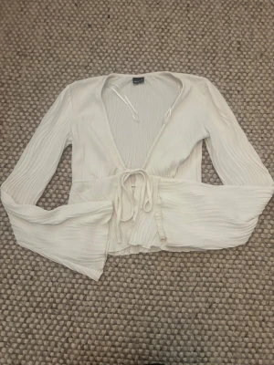 Vit omlottopp med knyt och vida ärmar - Säljer en superfin vit omlottopp med knyt i midjan och vida, plisserade ärmar. Toppen har en djup v-ringning och är gjord i ett lätt, luftigt material som känns perfekt för varmare dagar. Perfekt att styla med jeans eller kjol!