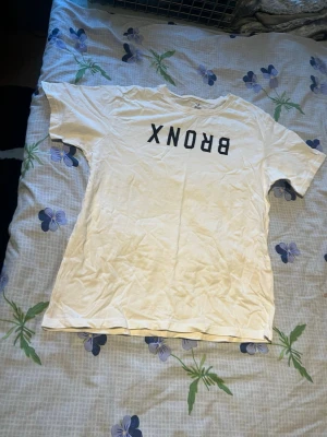 Vit t-shirt med BRONX-tryck - Vit t-shirt med svart text 'BRONX' på bröstet. Klassisk rak passform och rund halsringning. Perfekt för en avslappnad streetwear-look. Tillverkad i mjuk bomull och har korta ärmar.