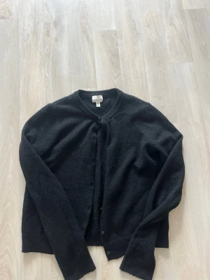 Svart kofta från H&M - Klassisk svart kofta från H&M med rund halsringning och knäppning framtill. Tillverkad i mjukt, stickat material som passar perfekt till lager på lager. Enkel och stilren design som funkar till det mesta.