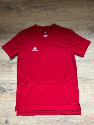 Röd sporttröja från Adidas - Snygg röd sporttröja från Adidas med vit logga på bröstet. Tröjan har V-ringning, korta ärmar och är tillverkad i ett lätt och snabbtorkande funktionsmaterial. Perfekt för träning eller match.