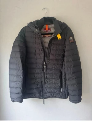 Svart pufferjacka från Parajumpers - Snygg svart pufferjacka från Parajumpers med huva och gul detalj vid halsen. Jackan har quiltad design, tvåvägsdragkedja och logotyp på ärmen. Perfekt för kalla dagar och riktigt skön att bära.