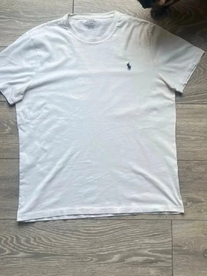 Vit t-shirt från Ralph Lauren - Klassisk vit t-shirt från Ralph Lauren med den ikoniska lilla hästloggan broderad på bröstet. T-shirten är kortärmad, har rund hals och är gjord i mjuk bomull för en skön känsla. Perfekt basplagg till garderoben.