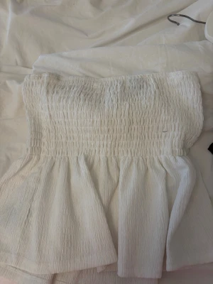 Vit bandeau topp från Gina Tricot - Säljer en vit bandeau topp från Gina Tricot i storlek M. Toppen är axelbandslös med smockad överdel och lätt utsvängd nederdel. Tillverkad i ett tunt, strukturerat bomullsliknande material som känns luftigt och perfekt för varma dagar.