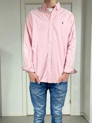 Ljusrosa skjorta från Polo Ralph Lauren - Säljer en ljusrosa skjorta från Polo Ralph Lauren med klassisk button-down krage och broderad logga på bröstet. Skjortan har långa ärmar och vita knappar framtill. Perfekt för dig som gillar stilrena och tidlösa plagg.