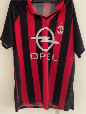 AC Milan matchtröja röd/svart - Säljer en klassisk AC Milan matchtröja i rött och svart med vertikala ränder. Tröjan har klubbmärke och Opel-logga på bröstet samt gul stjärna ovanför märket. Tillverkad i lätt och ventilerande material, perfekt för fotboll eller samling. En match tröja från sent 90 tal till tidigt 2000 tal 