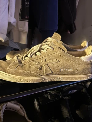Beige sneakers från Premiata - Stilrena beige sneakers från Premiata i mocka med vita detaljer och snörning. Skorna har en låg silhuett och coola mönster på sulan. Perfekta för dig som gillar en clean look.