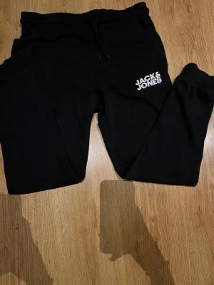 Svarta mjukisbyxor Jack & Jones - Svarta mjukisbyxor från Jack & Jones med vit logga på vänster lår. Byxorna har resår i midjan med dragsko och är tillverkade i mjukt material. Perfekta för chill eller träning.