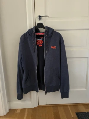 Blå zip hoodie från Superdry Nyskick - Mörkblå hoodie från Superdry med dragkedja, huva och röda detaljer på snörena. Klassisk logga broderad på bröstet. Två fickor framtill och ribbade muddar vid ärmar och nederkant. Perfekt för en chill och avslappnad stil. Knappt använd så den är som ny.