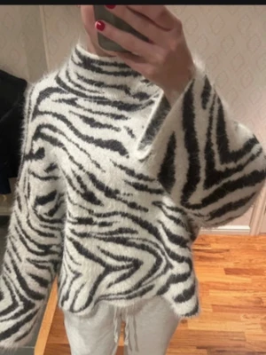 Zebramönstrad fluffig stickad tröja - Säljer en supermjuk stickad tröja med zebramönster i svart och vitt. Tröjan har hög krage, vida ärmar och en oversized passform. Perfekt för dig som vill sticka ut med ett djurmönster och gillar mysiga material. 