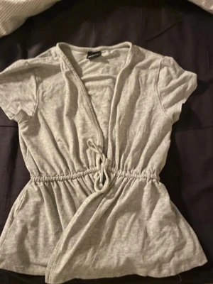 Grå omlottopp med knyt - Snygg grå omlottopp med korta ärmar och knyt i midjan. Toppen har en avslappnad passform och är tillverkad i mjuk bomull, perfekt för en chill och stilren look.