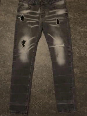 Gråa DSQUARED2 jeans - Säljer ett par grå jeans från Dsquared2 med slitningar och hål på benen. Jeansen har en tvättad look med vita detaljer och klassisk femficksmodell. Perfekta för dig som gillar en edgy och trendig stil.Har ett jättelitet hål i baksidan men kan enkelt sys ihop.
