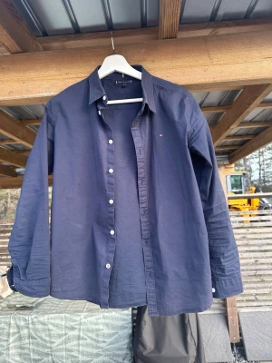 Mörkblå skjorta från Tommy Hilfiger - Klassisk mörkblå skjorta från Tommy Hilfiger med diskret logga på bröstet. Skjortan har lång ärm, knappar framtill och är tillverkad i bomull och elastisk för stretch. Barnstorlek 164. 