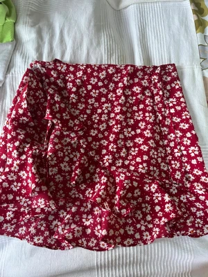 Röd blommig omlottkjol - Supersöt röd kortkjol med vita blommor och volangkant. Kjolen har omlottdesign som ger en snygg och avslappnad look. Perfekt för varma dagar och enkel att matcha med en enkel topp.för barn 11 år